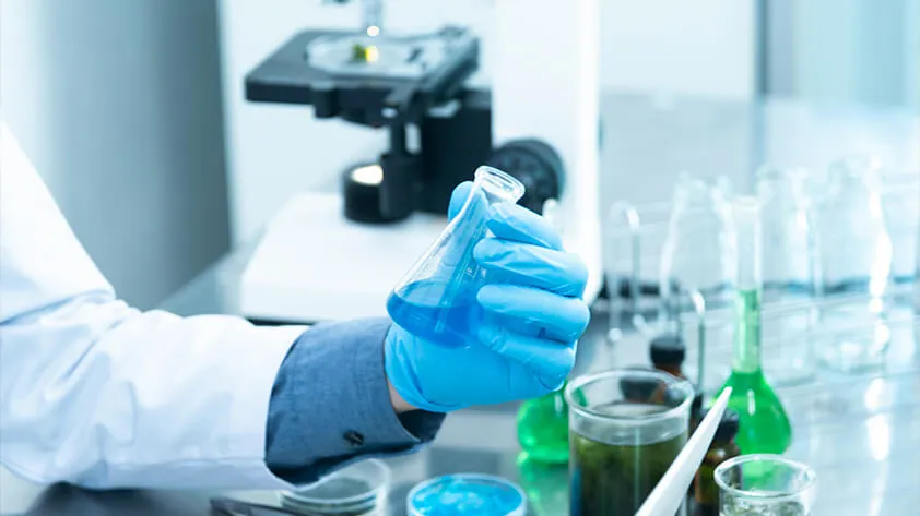 A researcher in a laboratory holding a flask of blue chemical liquid amidst scientific glassware and equipment