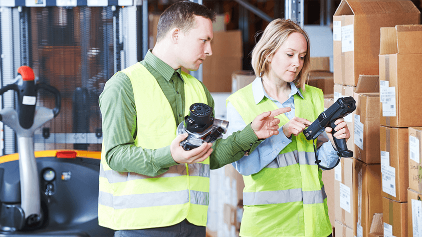 RFID Global Regulations A warehouse supervisor holding a handheld scanning terminal while guiding a colleague amidst stacked inventory boxes in a modern logistics facility