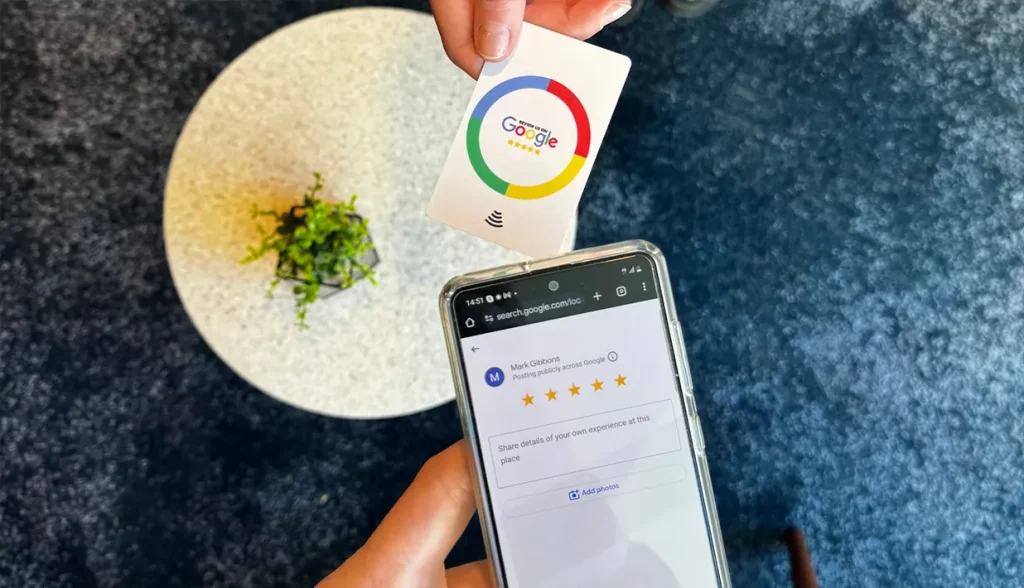 A person is tapping a white NFC Google Review card on the back of a smartphone. The phone screen shows the Google review interface, ready for a customer to leave a five-star rating.
