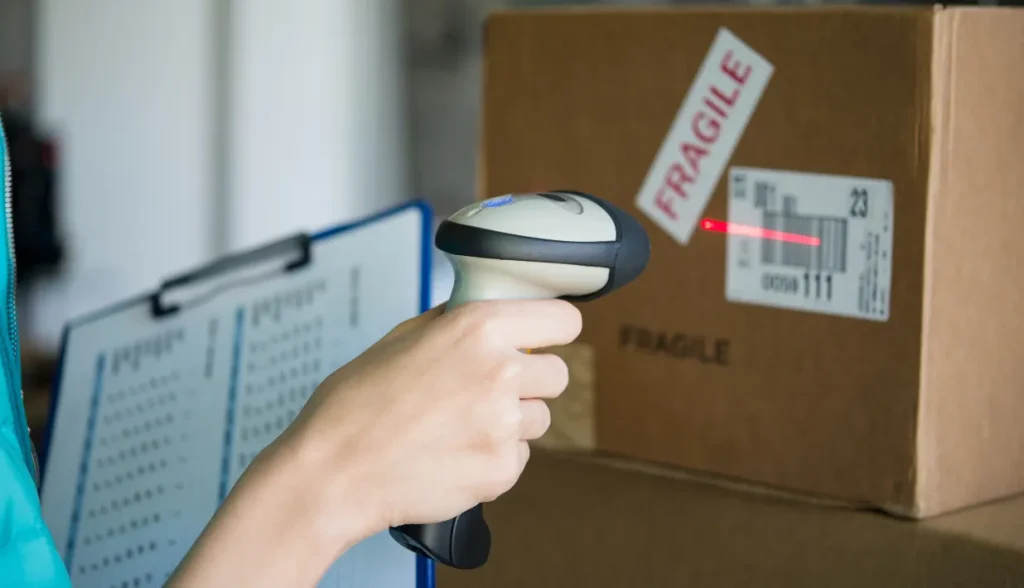 Hand holding a traditional barcode scanner, scanning a fragile-labeled box with a paper checklist in the background