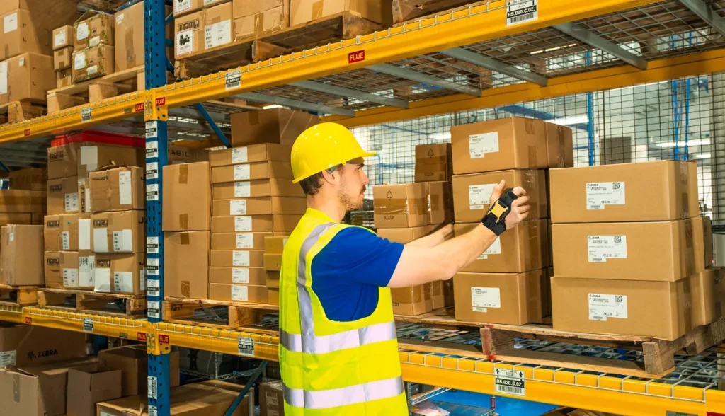 Warehouse worker in a hard hat and vest using a wearable RFID reader to scan stacked boxes on industrial shelving