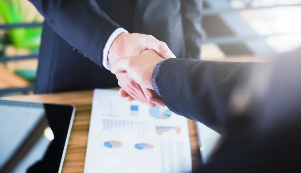 Two businesspeople shaking hands over a table with charts and a tablet.