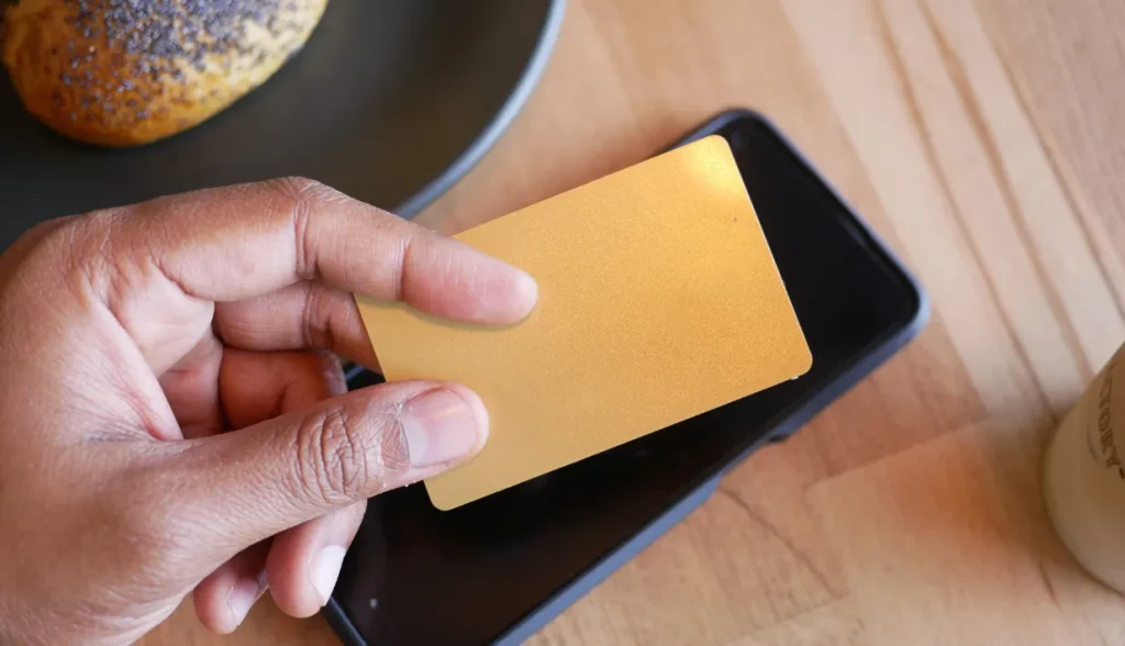 Hand holding a gold card near a smartphone, testing NFC’s reading distance.