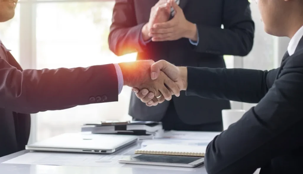 Business professionals shaking hands with a colleague clapping in the background.