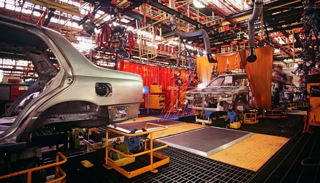 Automotive manufacturing plant floor with vehicle frames on an assembly line and welding equipment in operation.