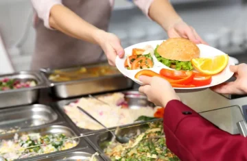 A cafeteria worker hands a plate of food to a student in a school dining hall, with salad bars in the background.