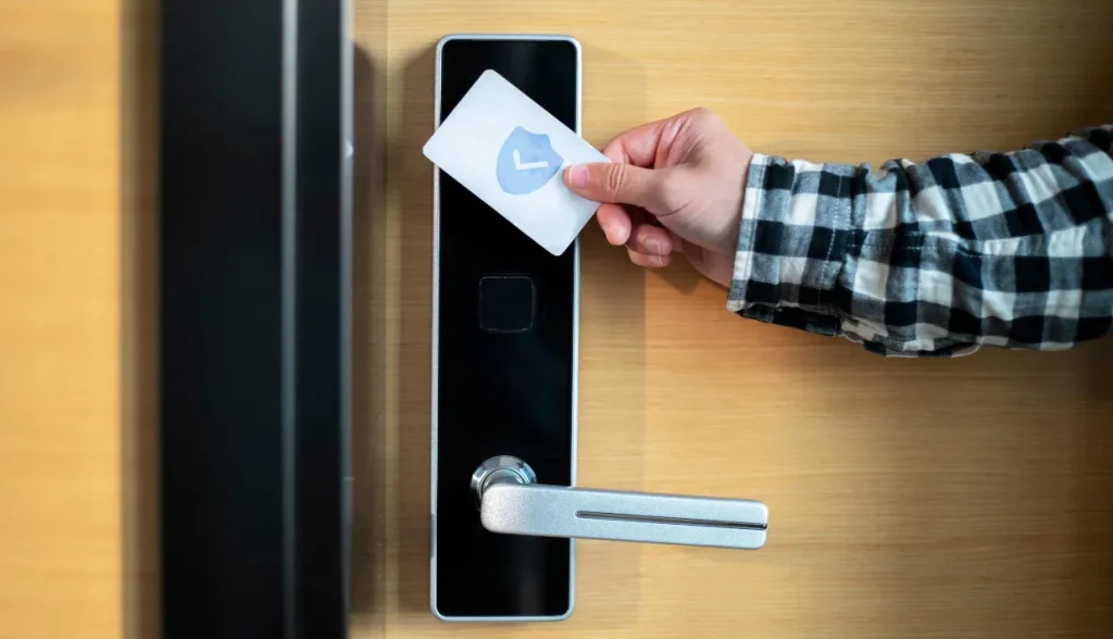 A hand holding a secure RFID key card with a shield icon up to a modern door lock.