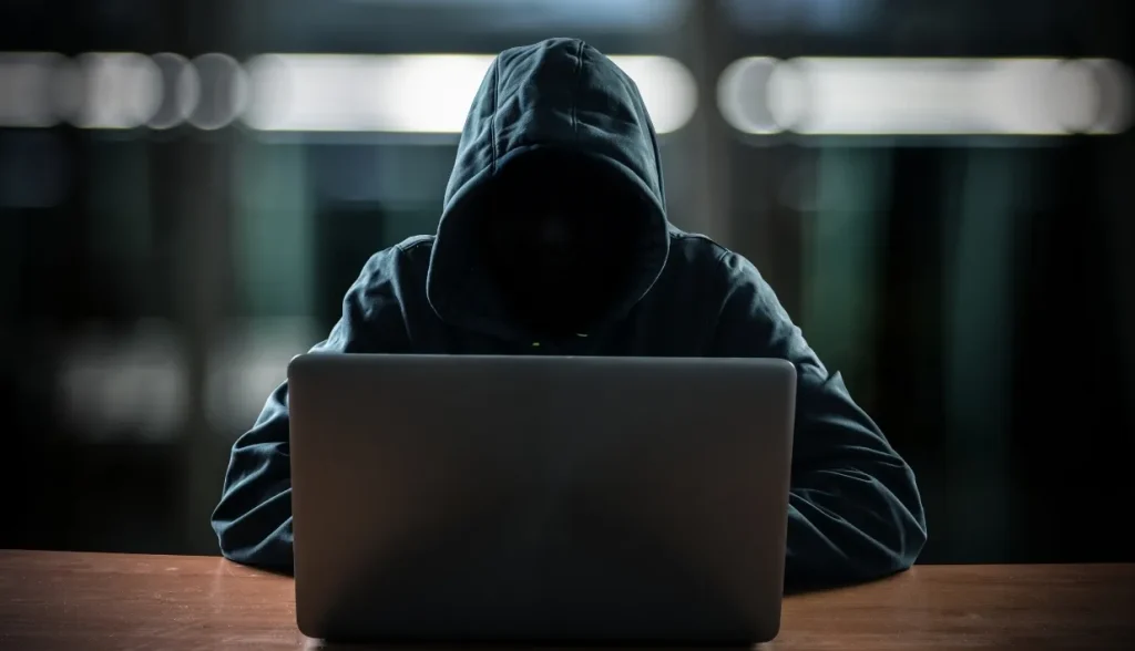 A hooded individual using a laptop in a dim setting, representing cyber threats to NFC payments like skimming or eavesdropping.