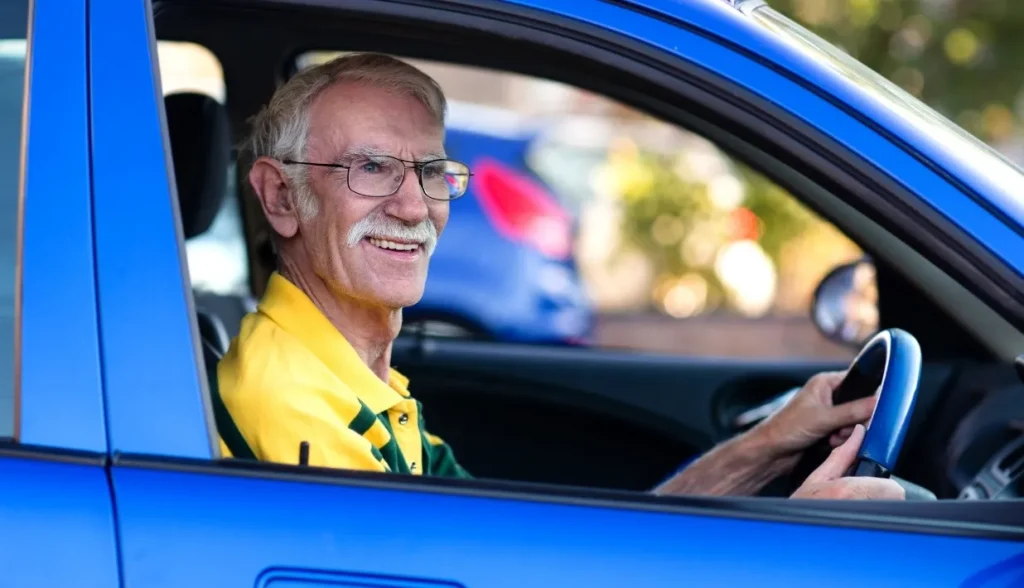 Older man with white hair and mustache driving a blue electric car, smiling at the camera.