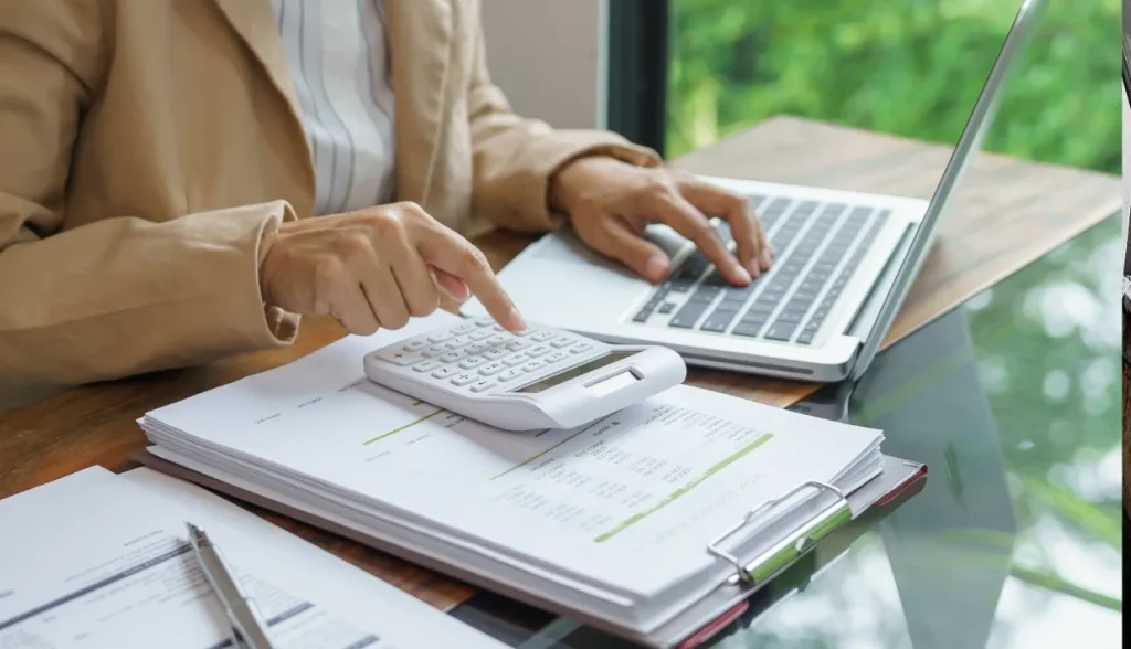 Person in a beige blazer using a calculator and laptop on a desk with financial documents, analyzing data.